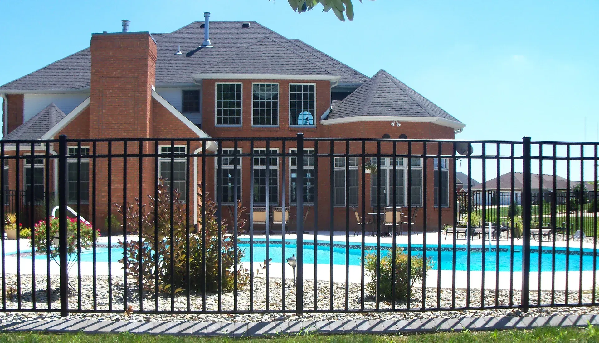 Black metal fence in front of a large brick house with a pool on a sunny day.