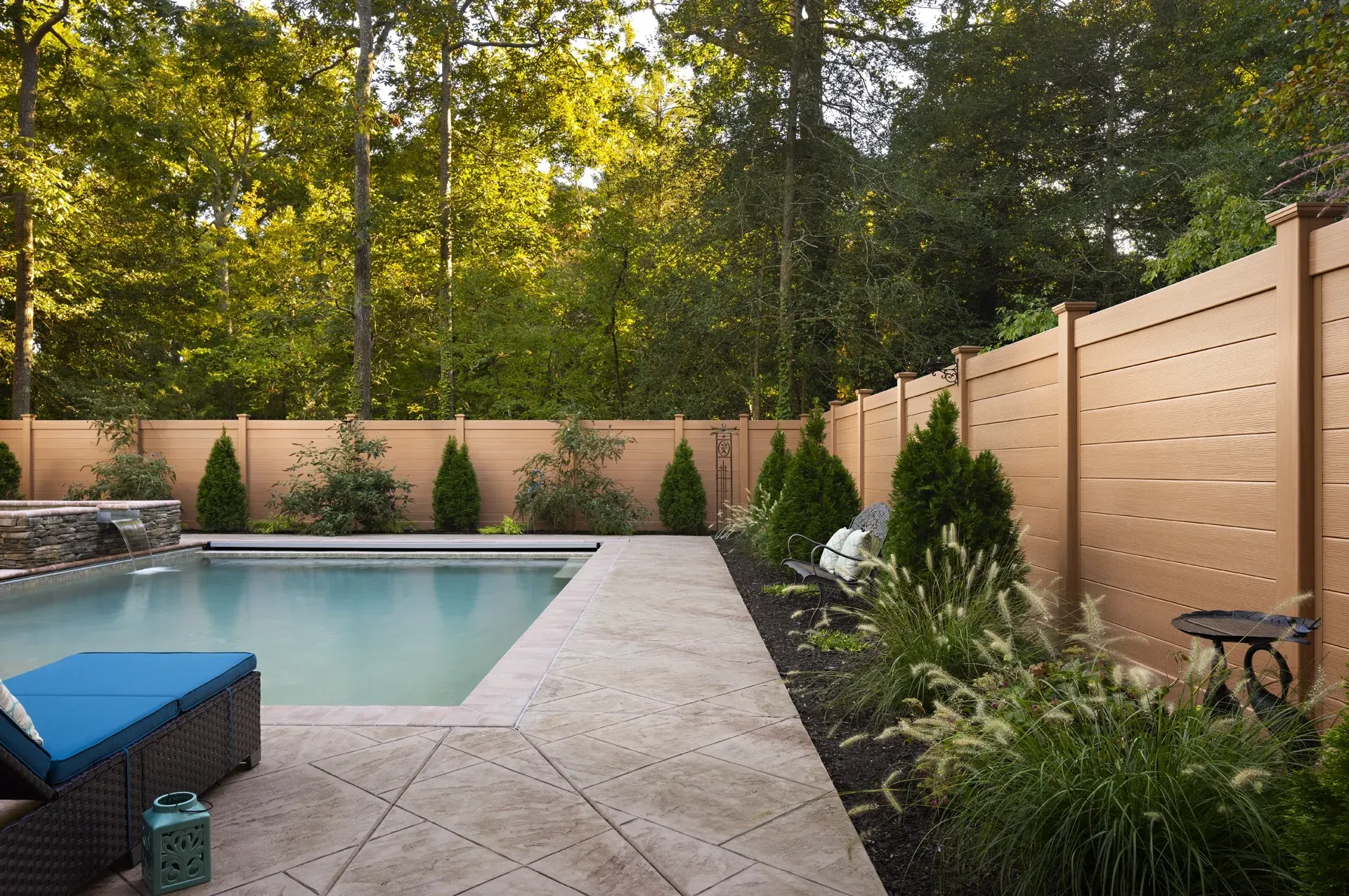 Poolside view with brown fence, trees, and greenery. A blue lounge chair sits by the pool.