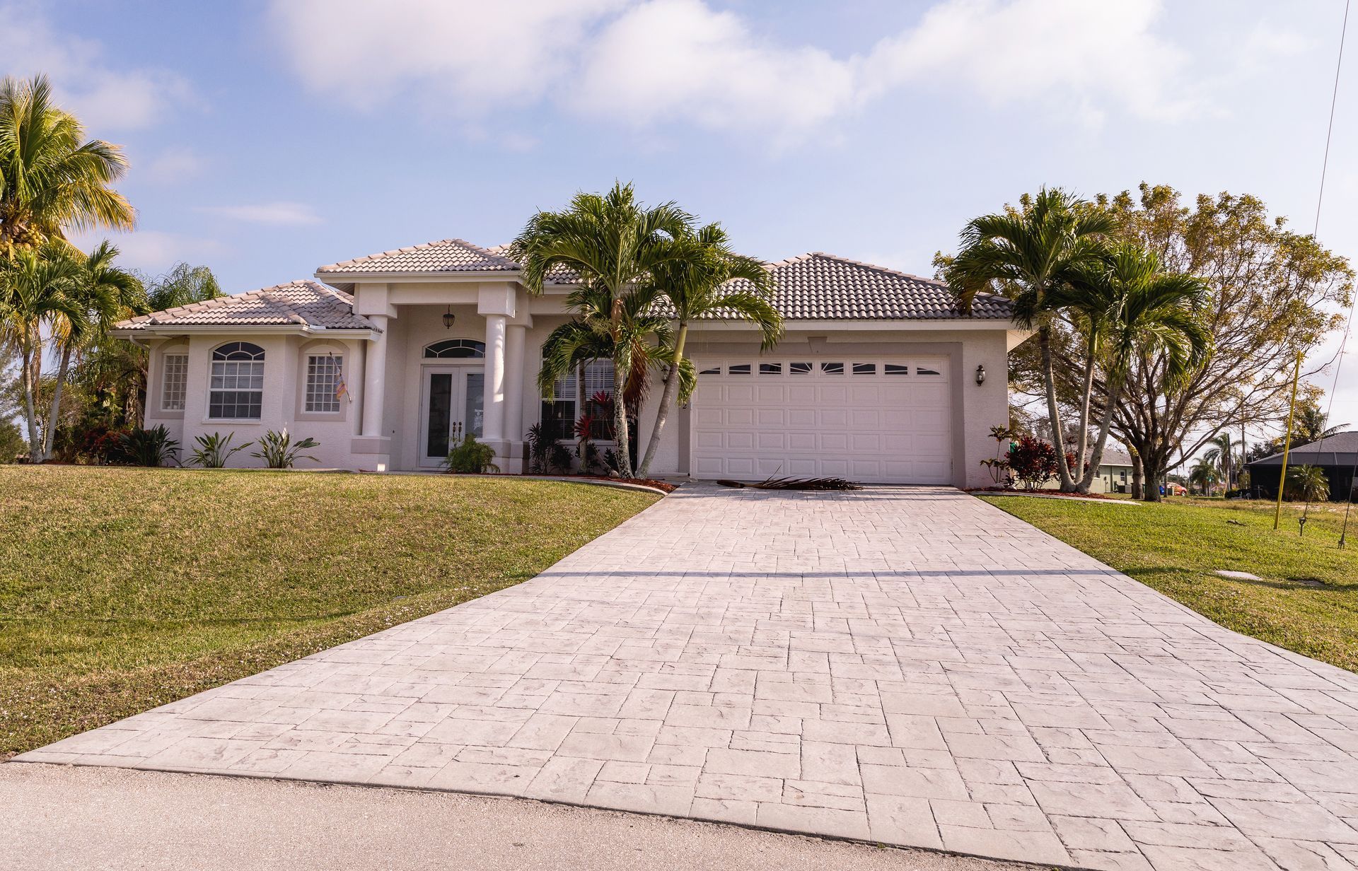 White suburban home with a gray brick-patterned driveway, palm trees, and a blue sky.