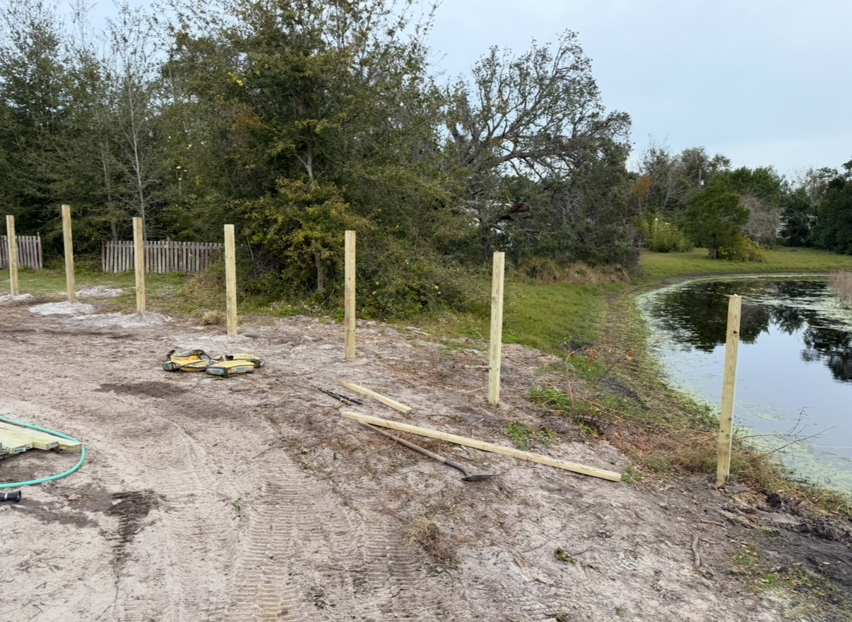 Wooden fence posts installed along a pond's edge on a dirt ground, with trees in the background.