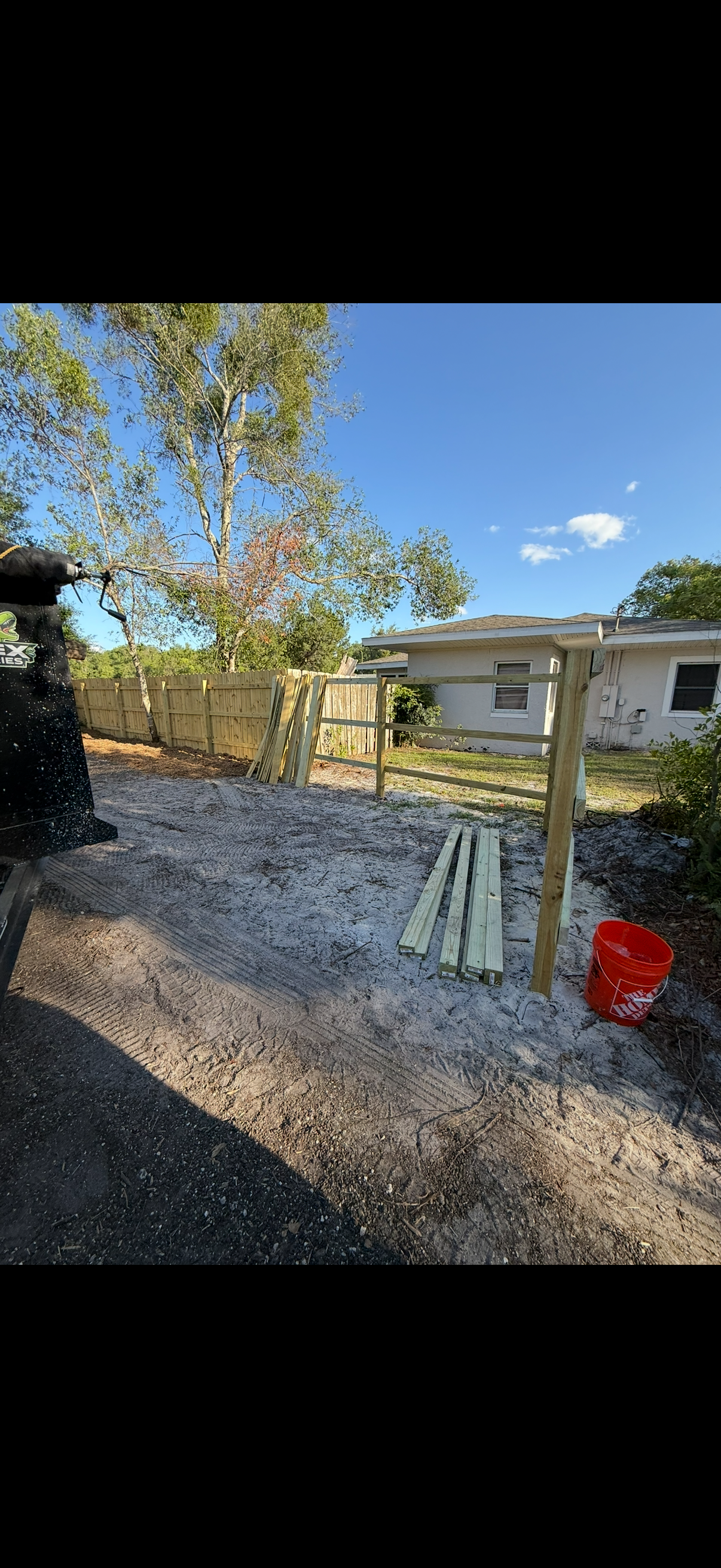 Construction of a wooden fence with posts, gravel ground, and building materials; trees and a house in the background.