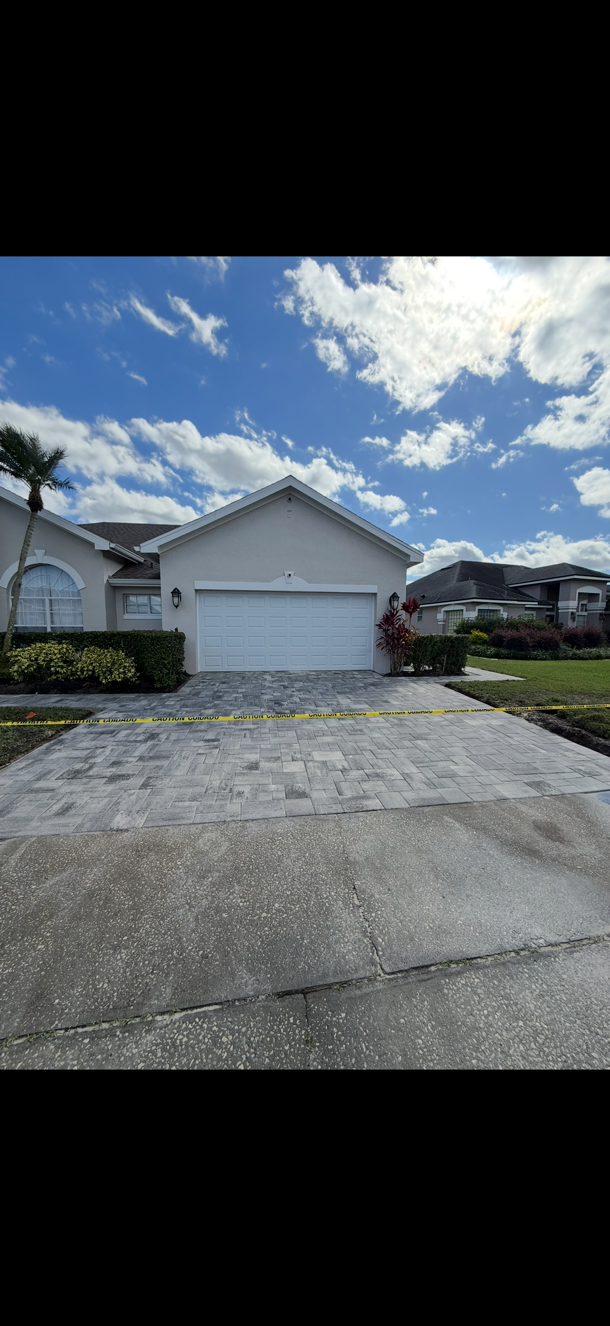 A house with a white garage door sits behind a cracked driveway and yellow caution tape under a cloudy sky.