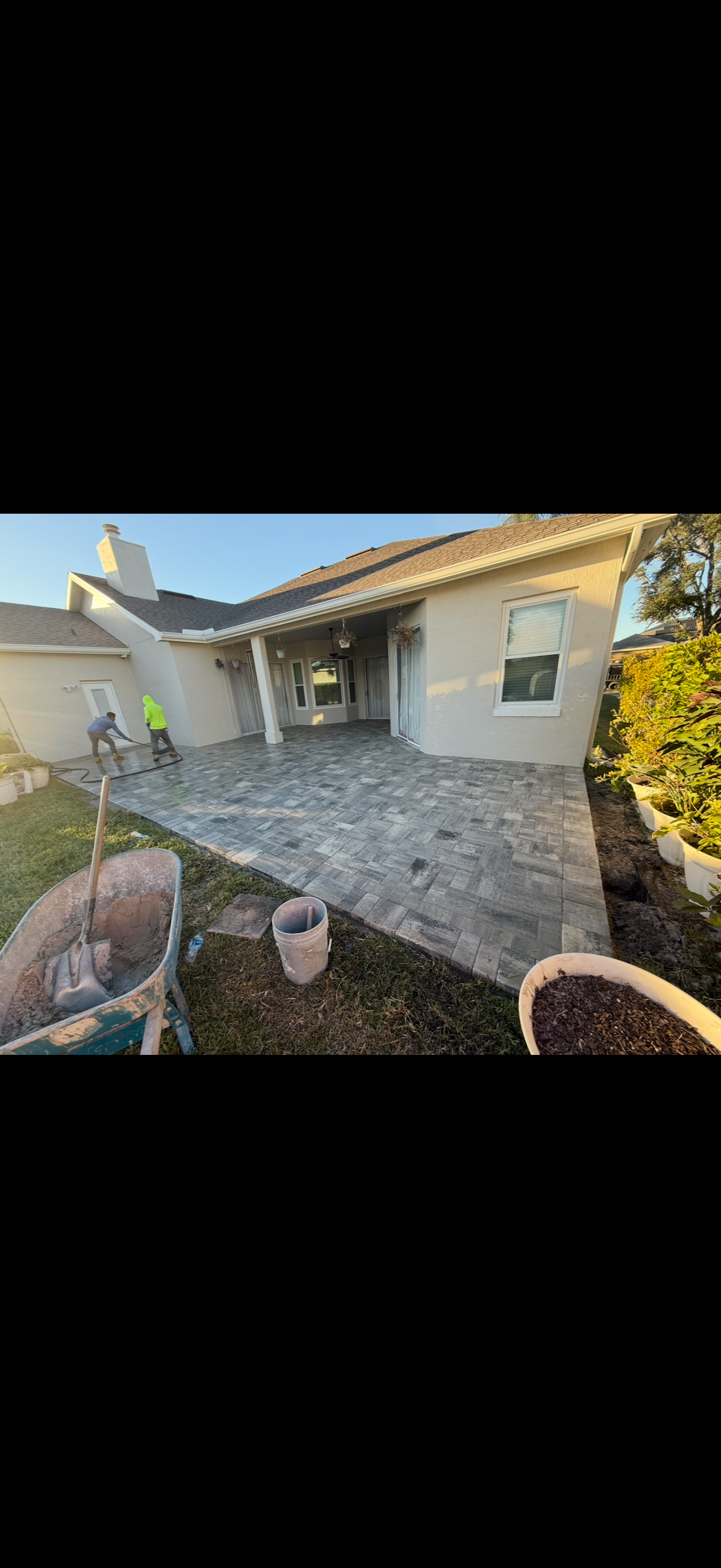 A backyard patio construction scene. A worker in safety vest near a house, with pavers and pots nearby.