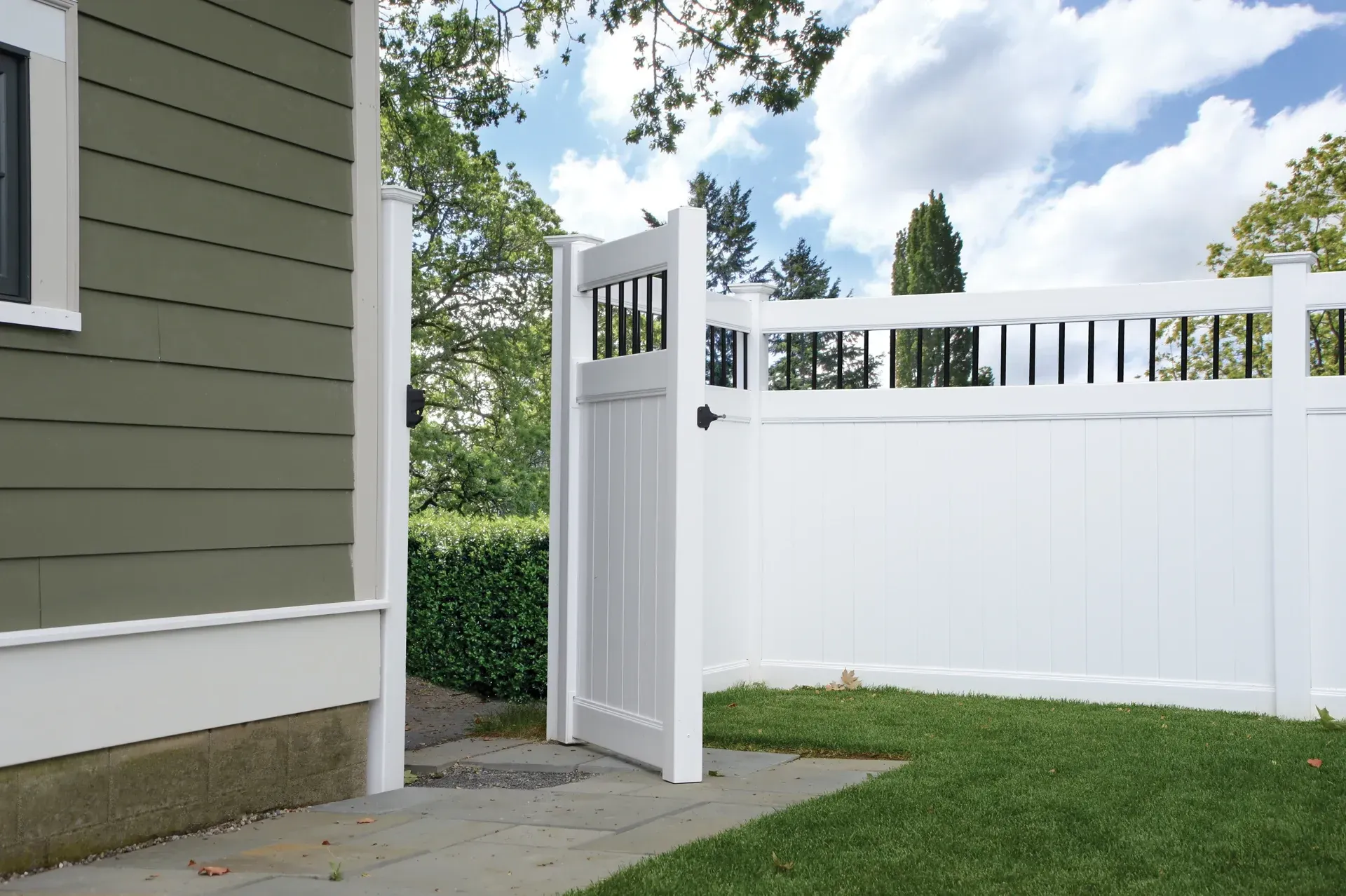 White fence and gate enclosing a grassy yard. A green house is on the left. Blue sky in background.