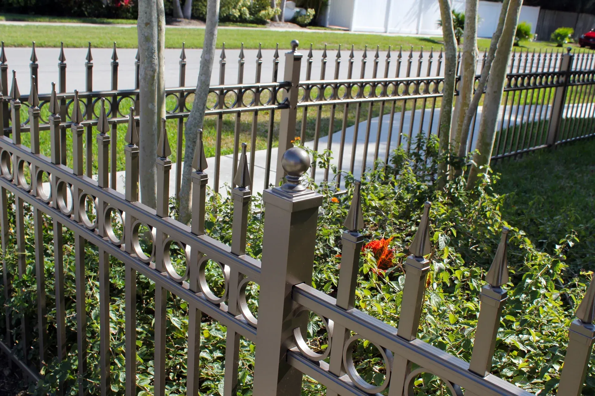 Brown metal fence with decorative circles and pointed finials encloses a green lawn, sidewalk, and trees.
