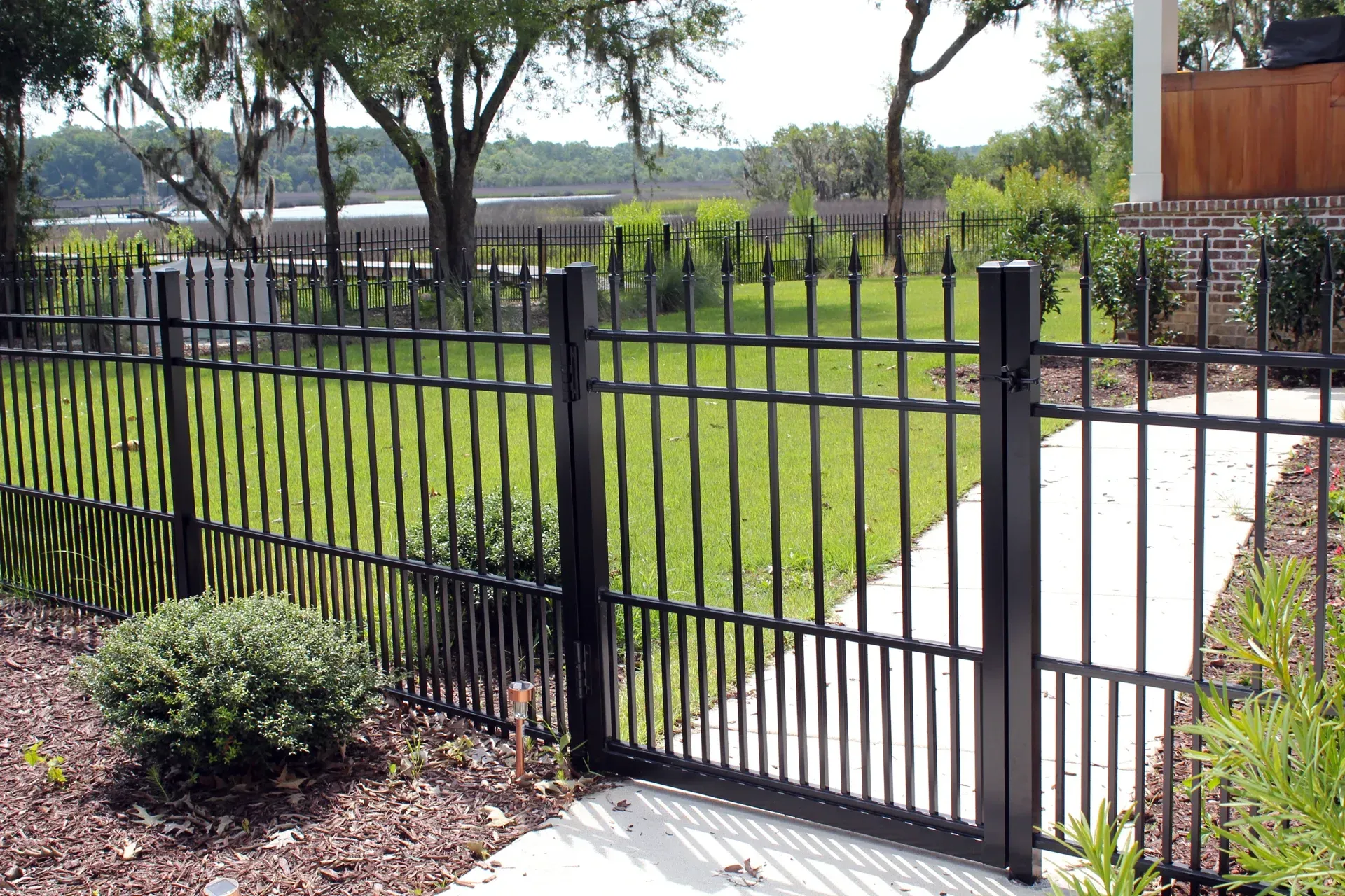 Black metal fence with gate in a grassy yard, surrounded by plants and trees.