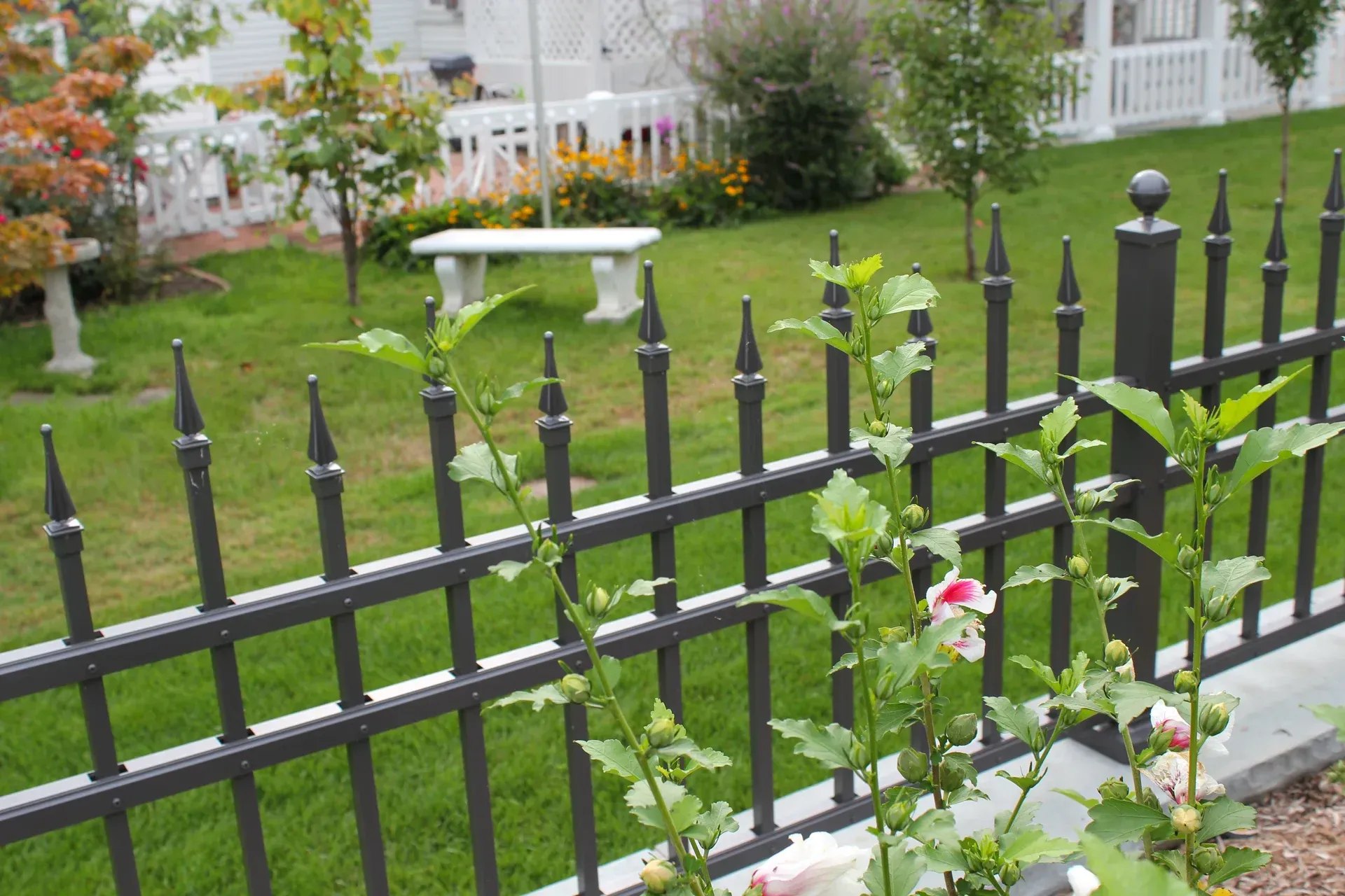 Black metal fence in front of a green lawn with a white bench and flowers.