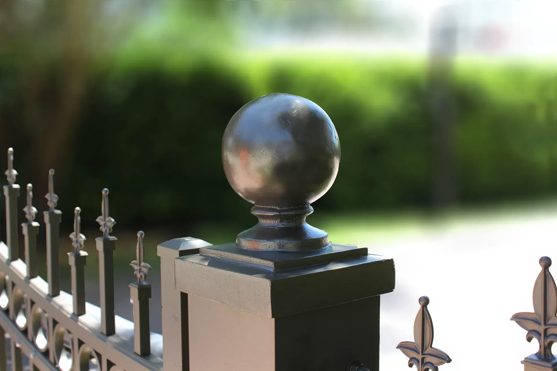 Black metal gate post topped with a sphere, in front of a blurred green hedge.