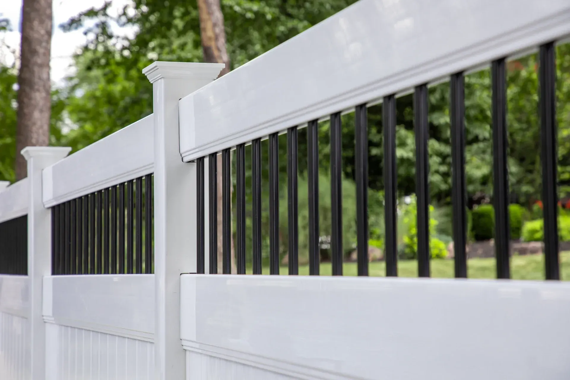 White and black vinyl fence with black vertical balusters, outdoors with green trees in background.