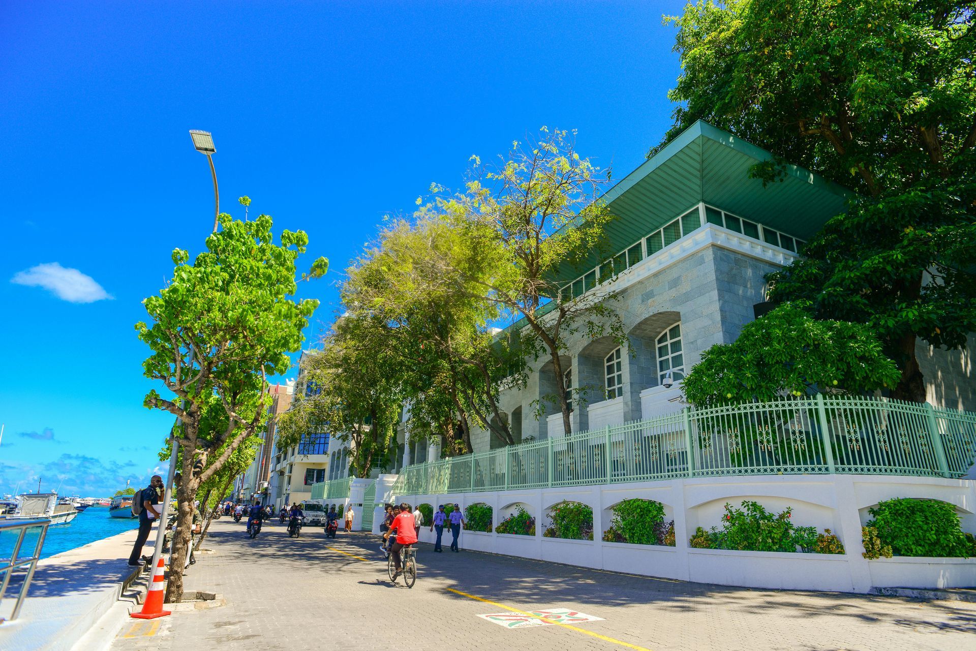 Street scene along a waterfront with a building, people walking and biking under a blue sky.