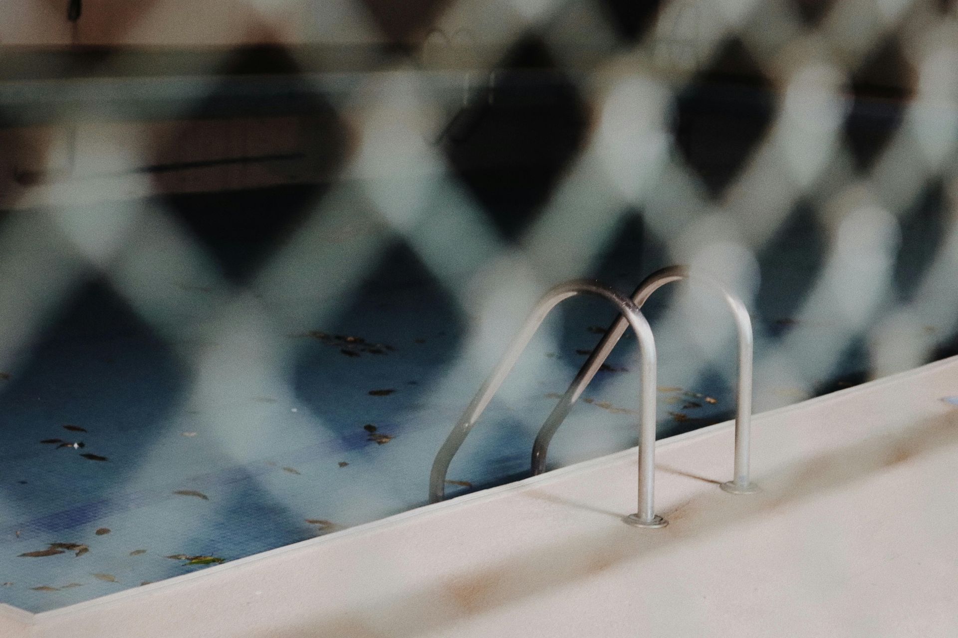 Pool with ladder, viewed through a chain link fence. Blue water, white concrete deck, shadows cast.