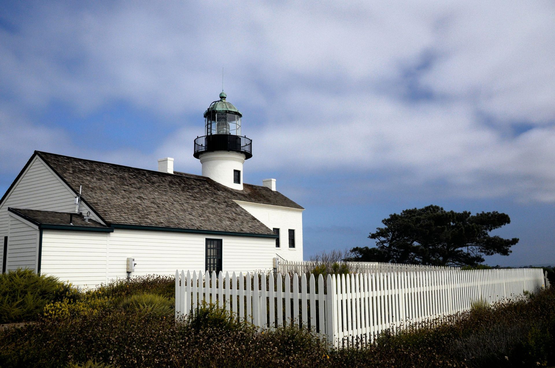 White lighthouse with black top, green dome, and white picket fence against a blue sky with clouds.