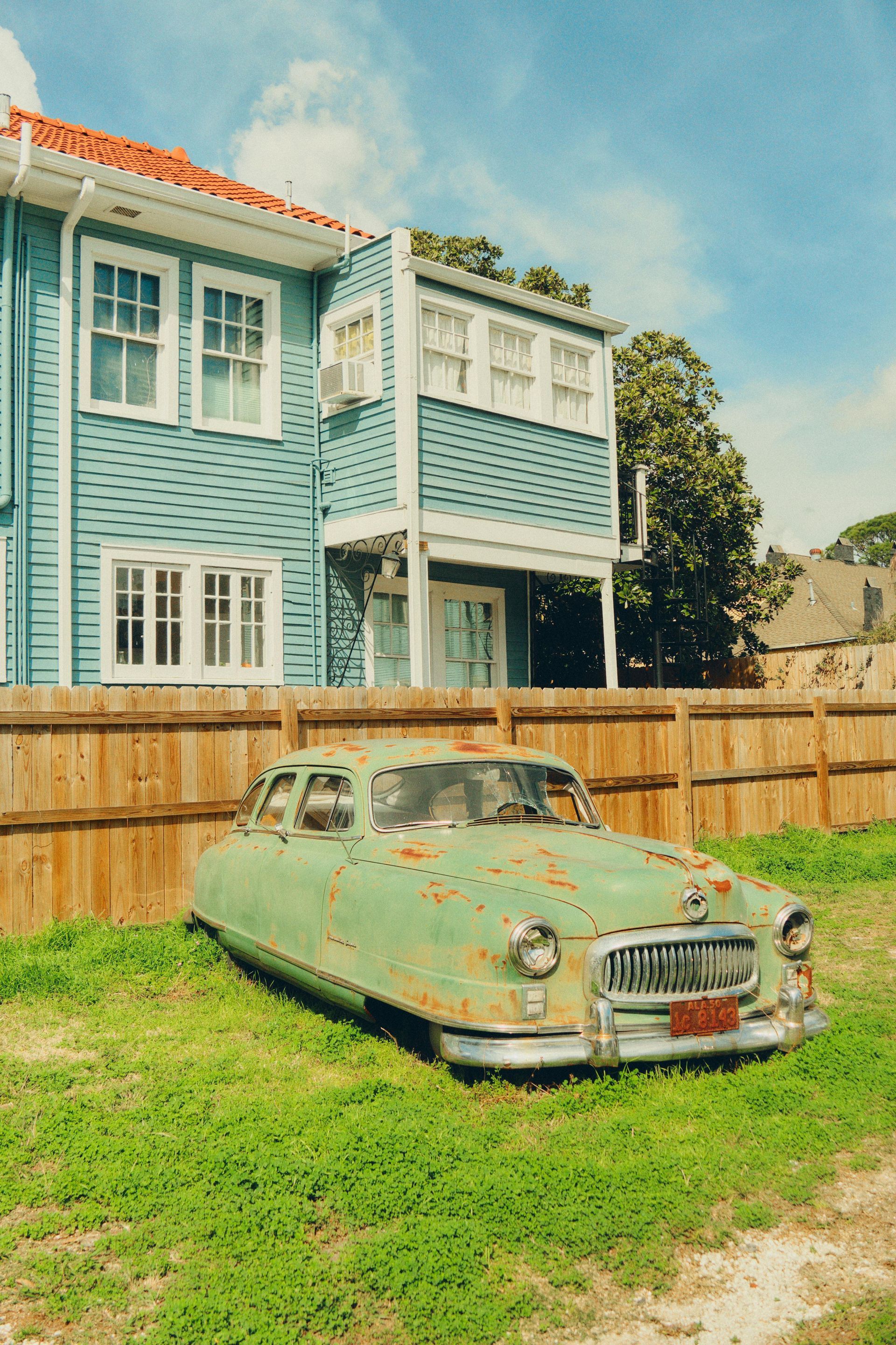 Green, rusty vintage car parked in front of a blue house with a wooden fence.