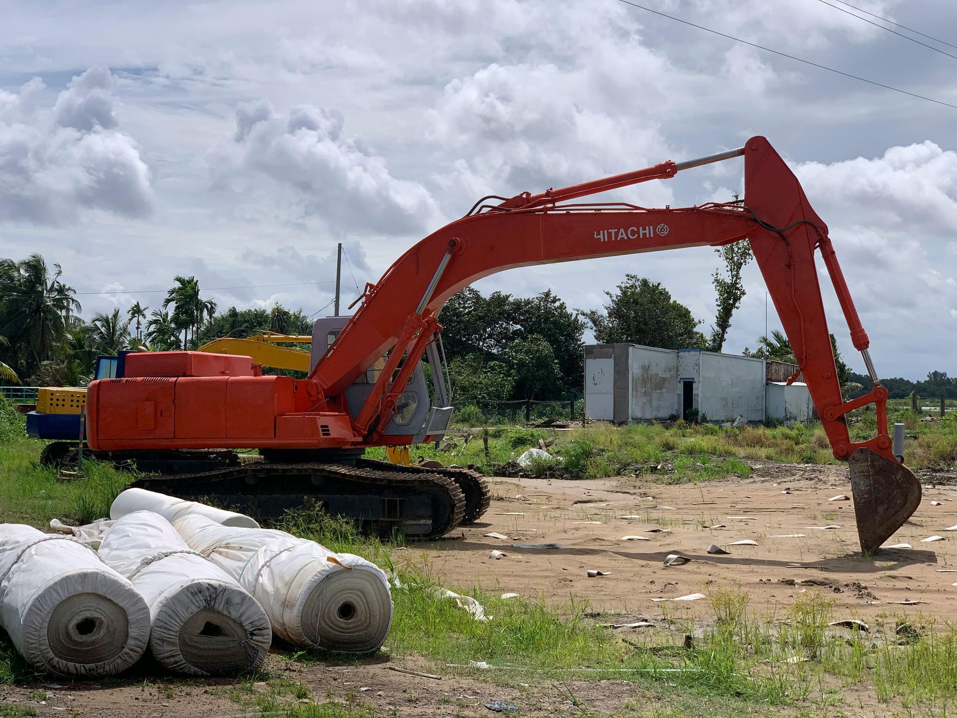 Red excavator on a construction site with rolls of white material in front. Overcast sky.