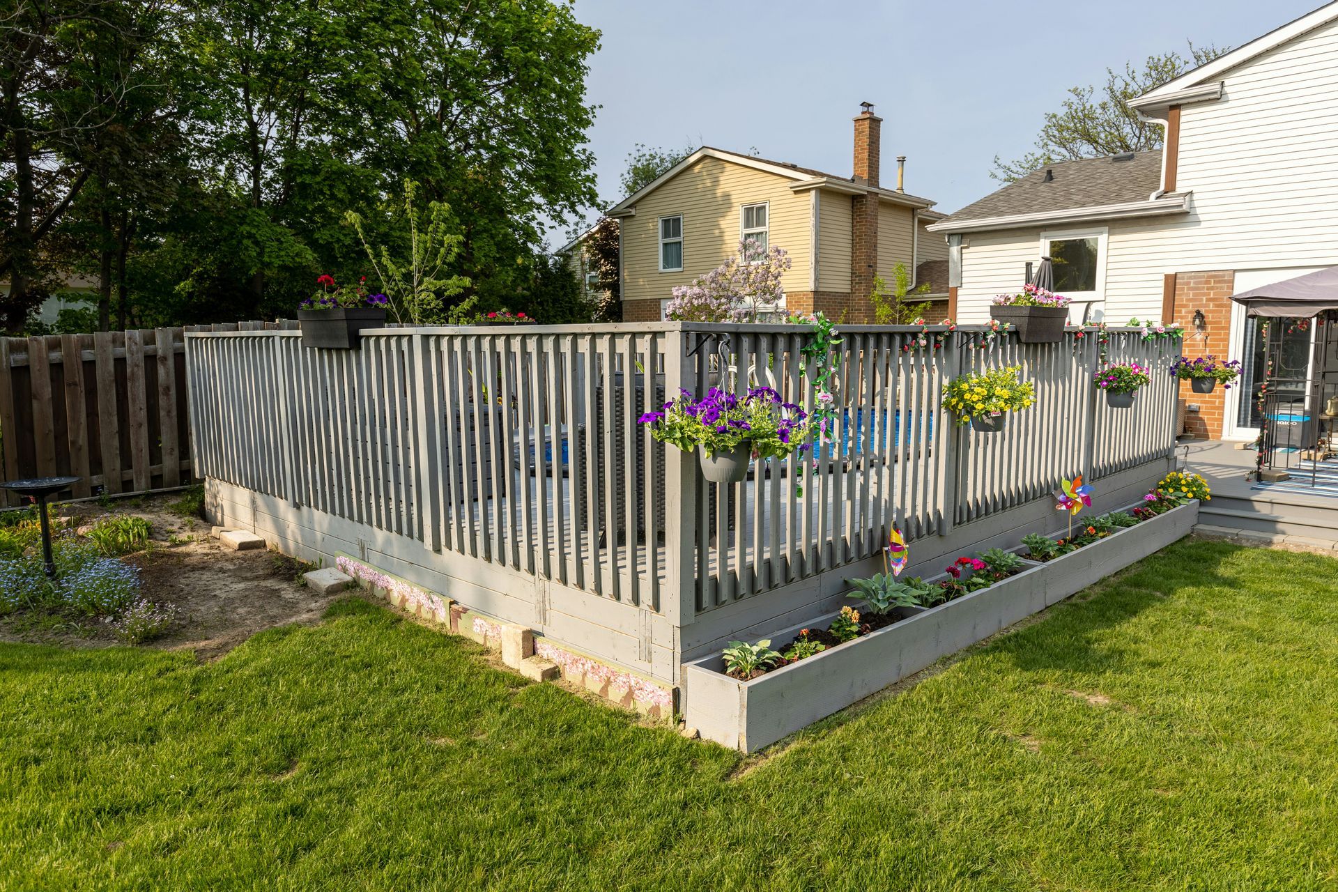 Gray wooden deck with flower boxes and potted plants in a backyard with green grass and houses.