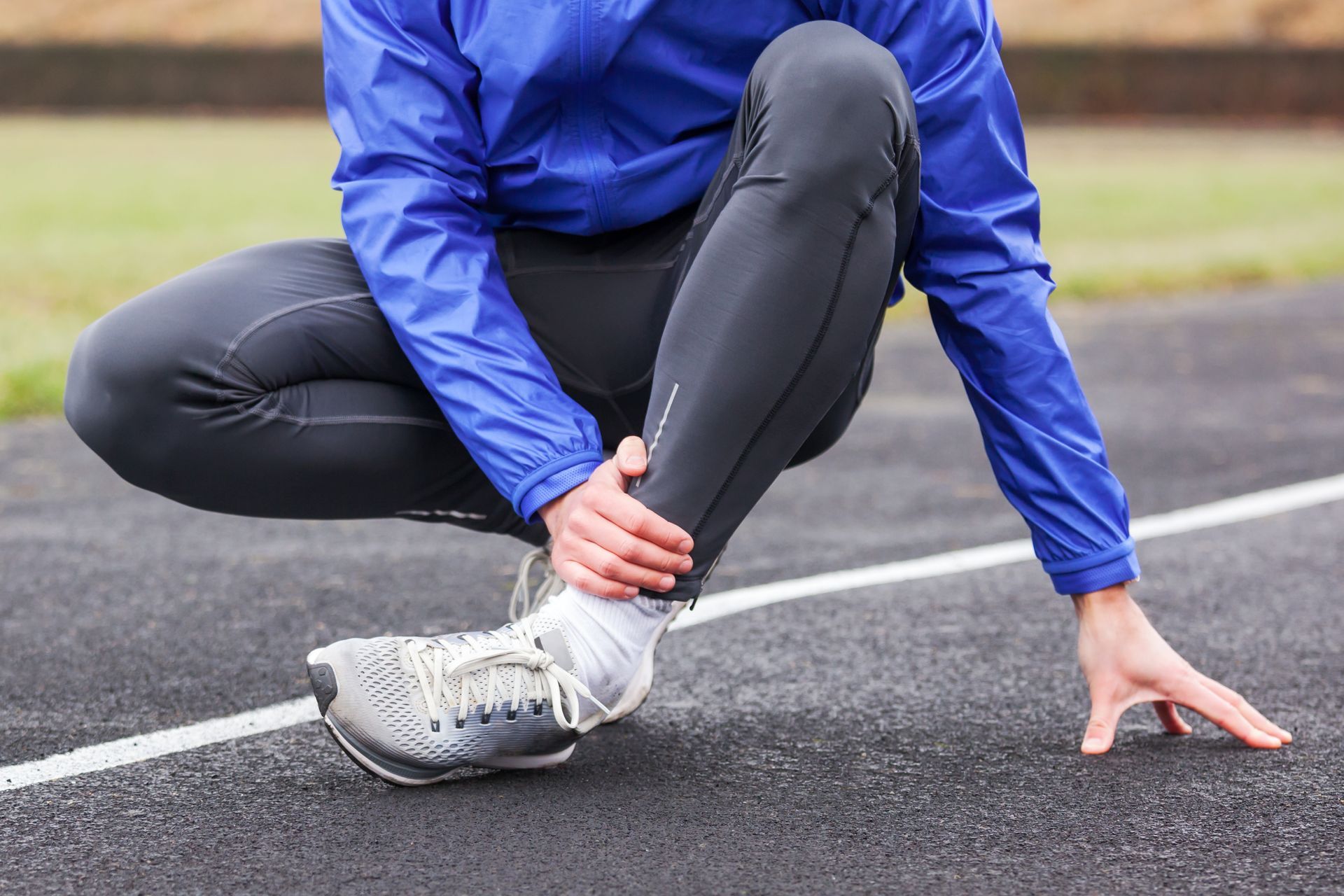 Runner on track, holding ankle, likely injured. Blue jacket, black leggings, gray shoes.