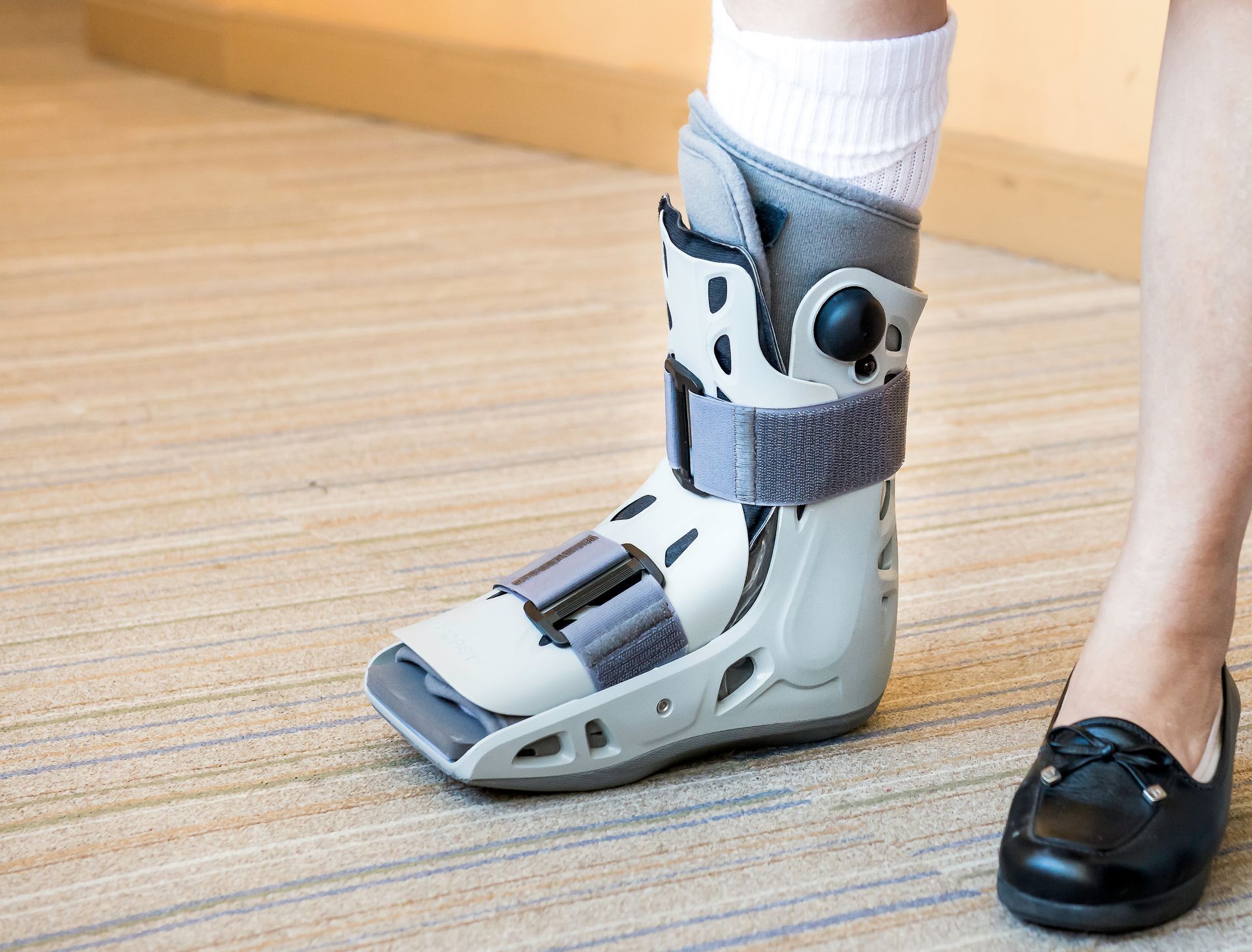 Person wearing a medical walking boot; white sock and black shoe visible. On a wooden floor.