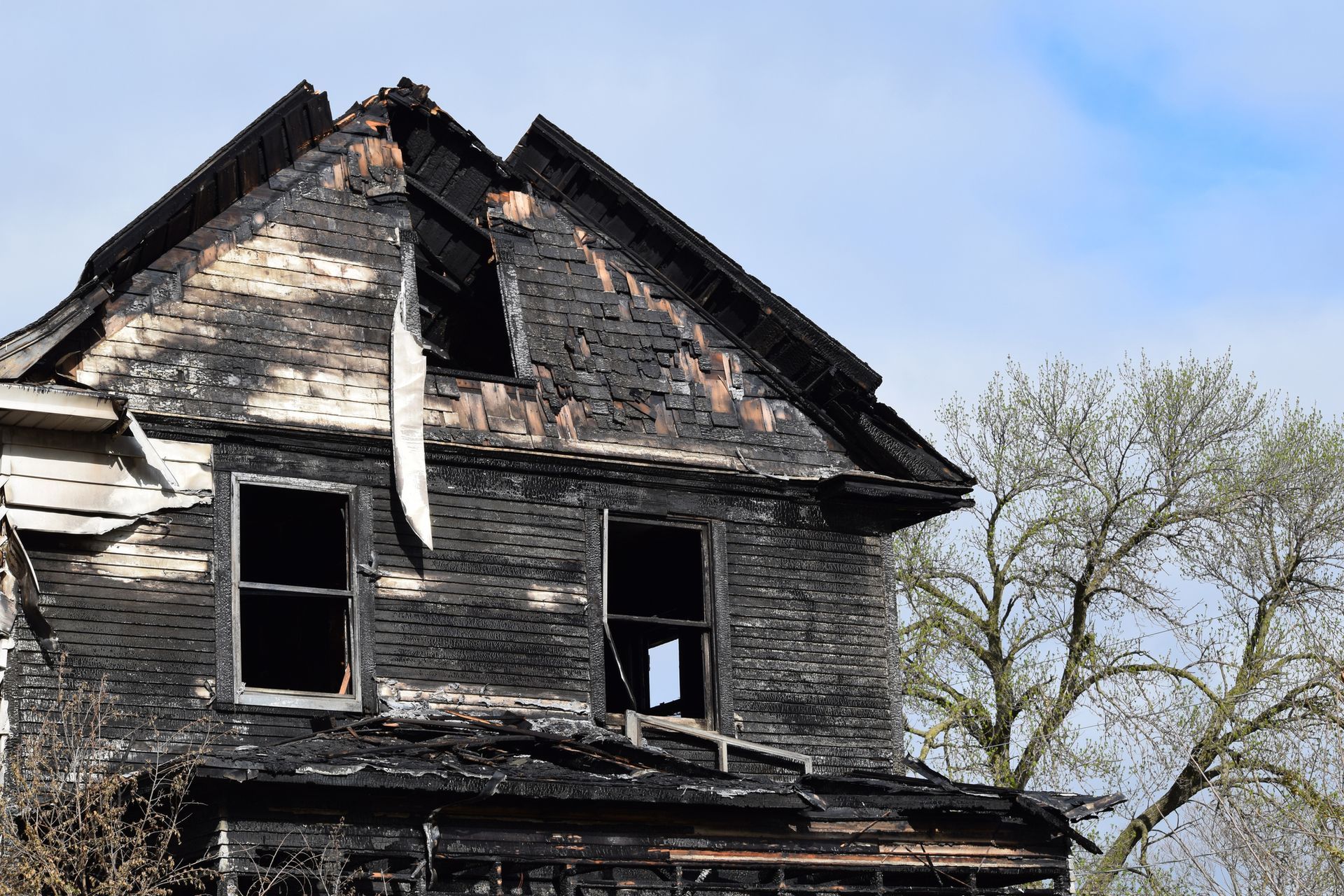 Burnt two-story house with black charred exterior and damaged roof; tree in the background.