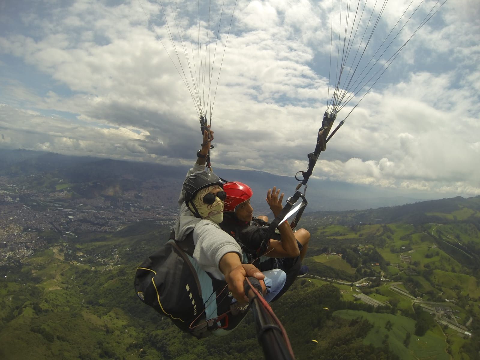 Dos personas haciendo parapente, tomándose un selfie con una ciudad y colinas al fondo.