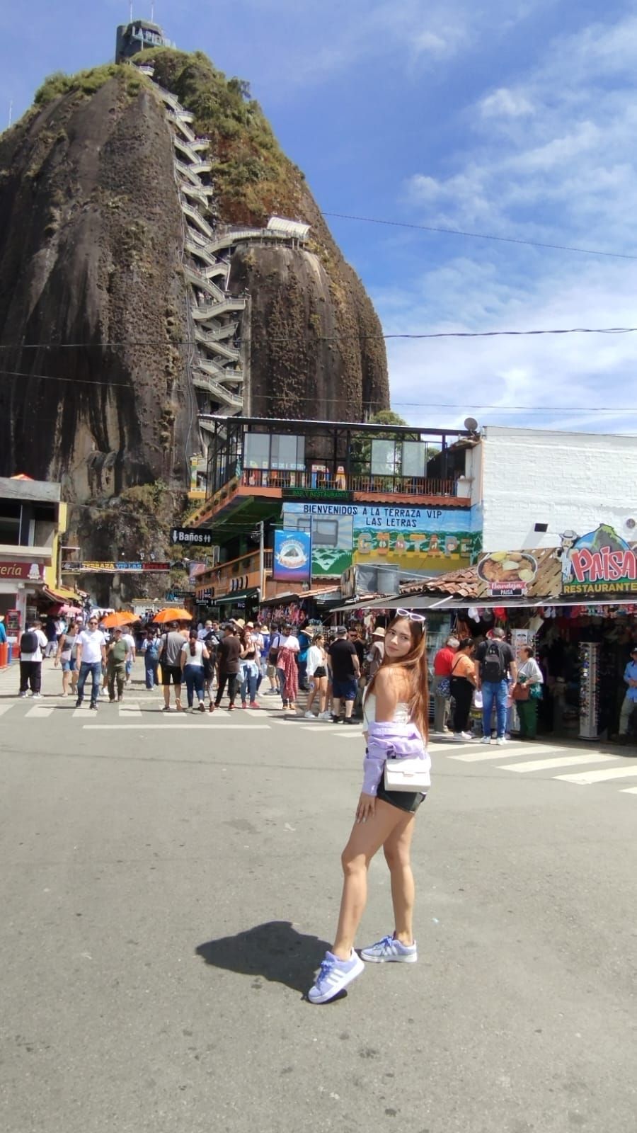 Una mujer está de pie frente a una gran formación rocosa con escaleras. Se ven gente y tiendas. Día soleado.
