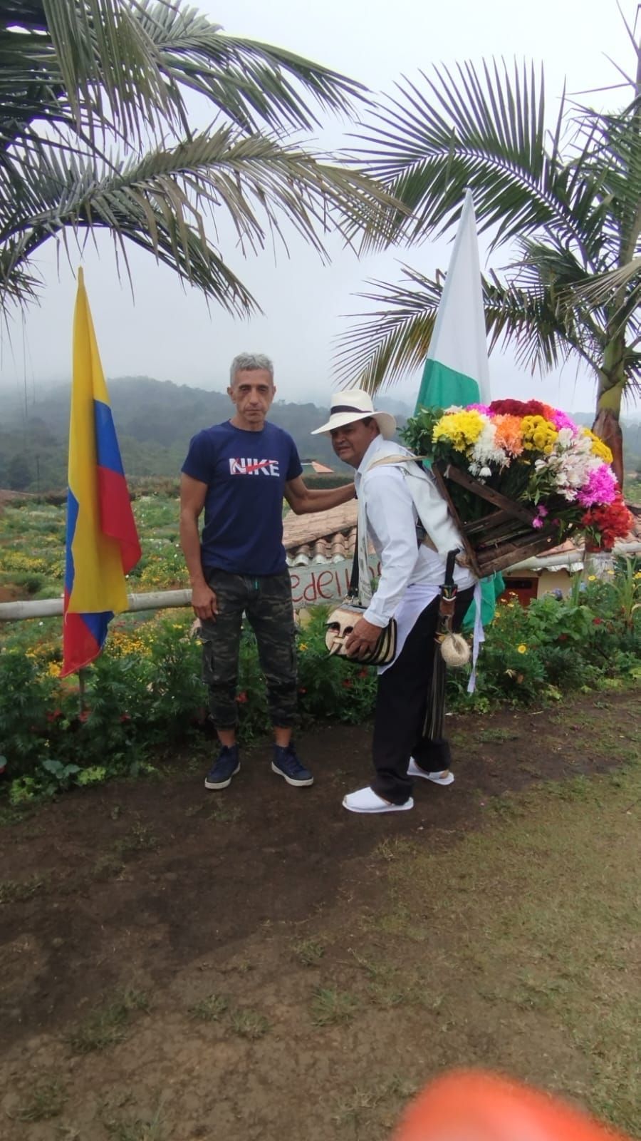 Hombre posa con vendedor de flores llevando cesta de flores; banderas colombianas en el fondo.