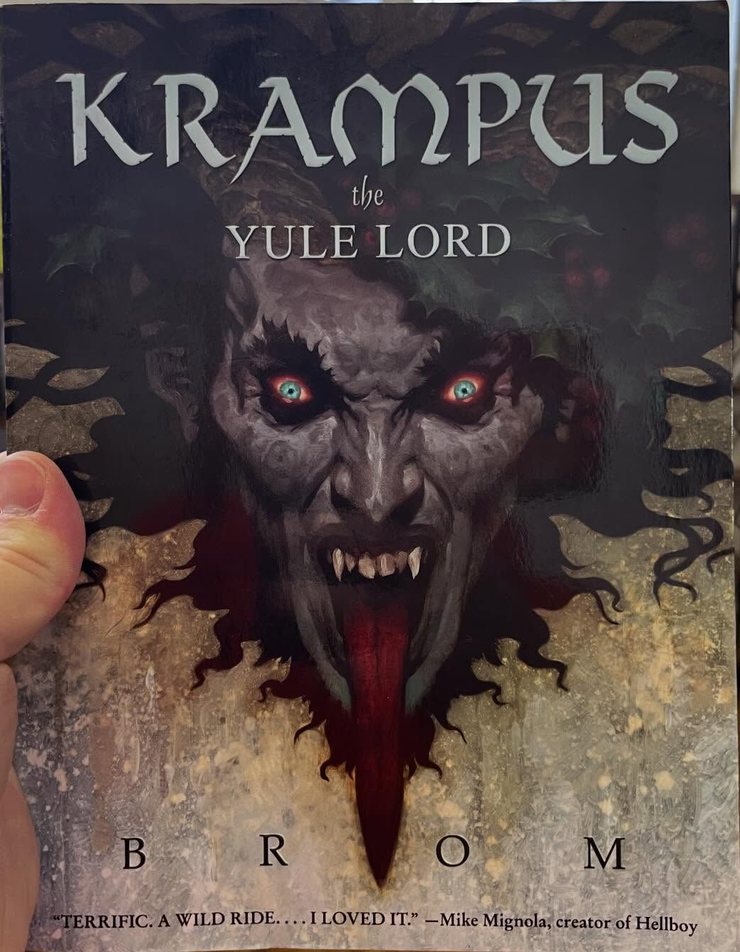 A hand holds a copy of the book "Krampus: The Yule Lord" by Brom in a shop, featuring a sinister, fanged creature on cover.