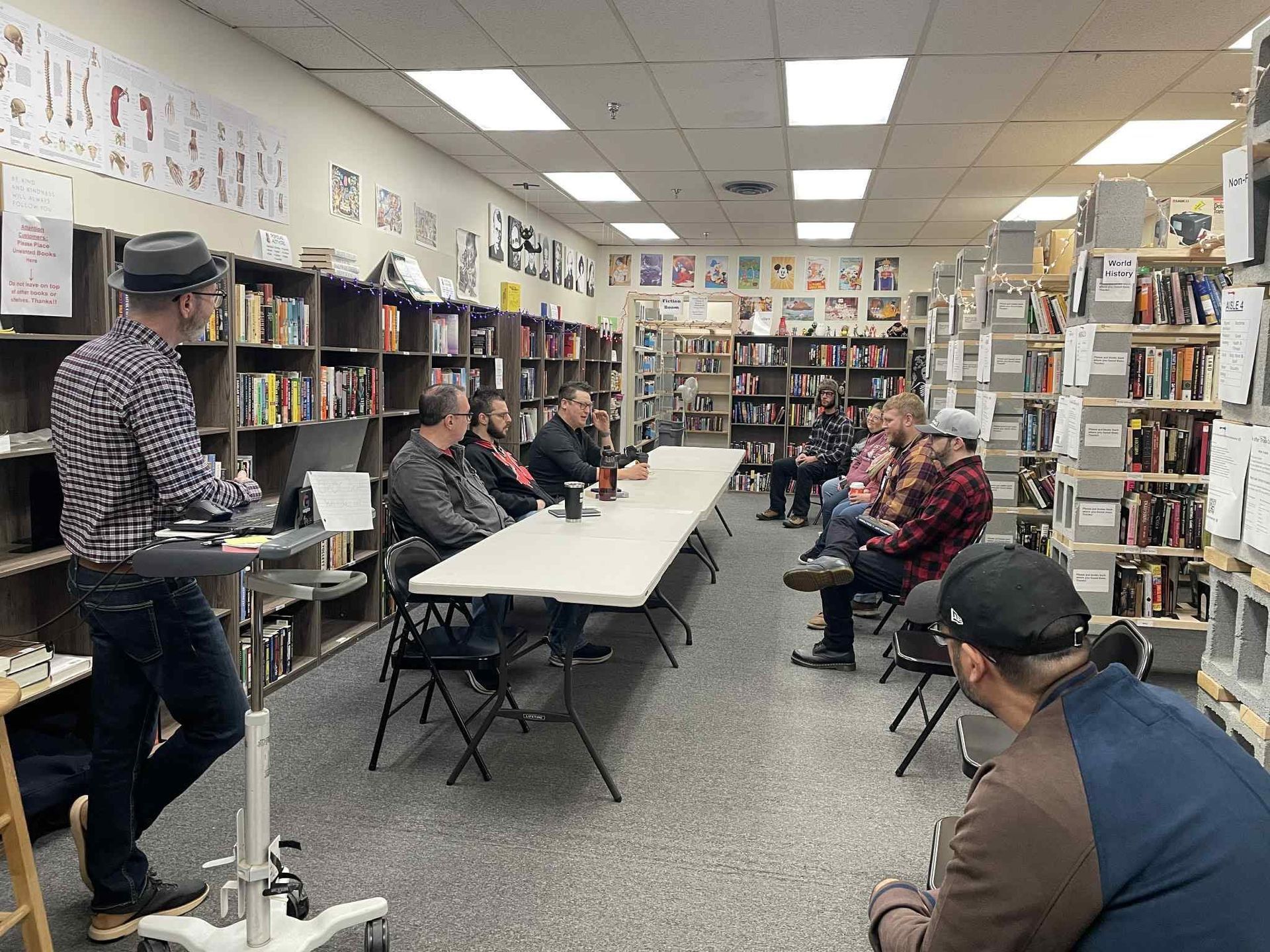 A speaker stands at a podium in a bookstore, addressing a group of people seated around a long table.