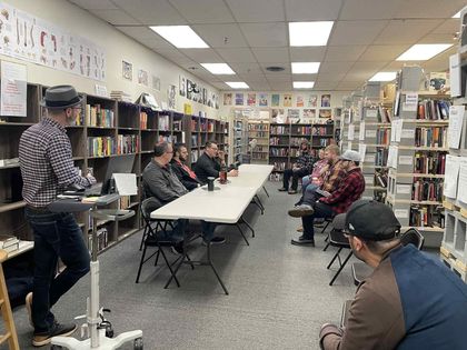 A speaker stands at a podium in a bookstore, addressing a group of people seated around a long table.