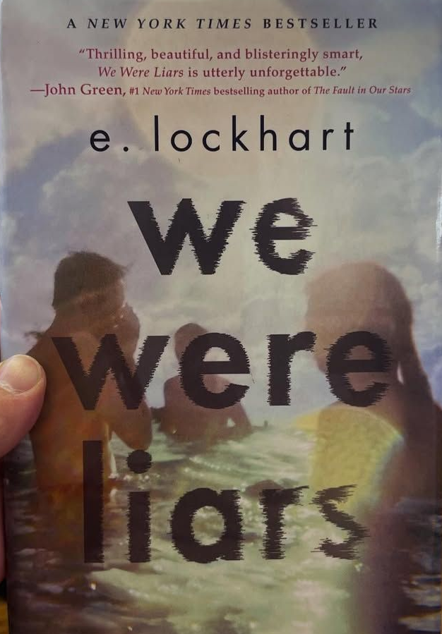 A hand holds the book "We Were Liars" by E. Lockhart in a room with shelves and a wooden floor.