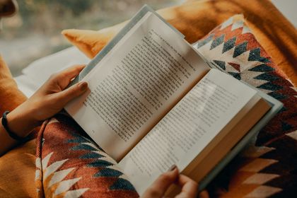 A person holding an open book, resting on a patterned, warm-toned textile.