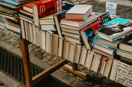 A wooden table displays a large collection of stacked and arranged books in an outdoor setting on a stone-paved surface.