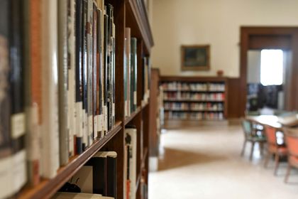 A close-up of a wooden library bookshelf with blurred books, tables, and chairs in the background.
