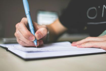 A close-up of a person writing in a notebook with a light blue pen on a neutral background.