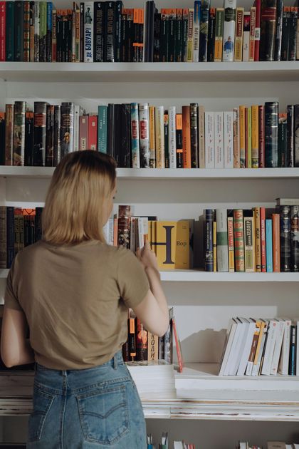 A person in a tan shirt and jeans standing before a tall bookshelf, reaching for a book on a middle shelf.