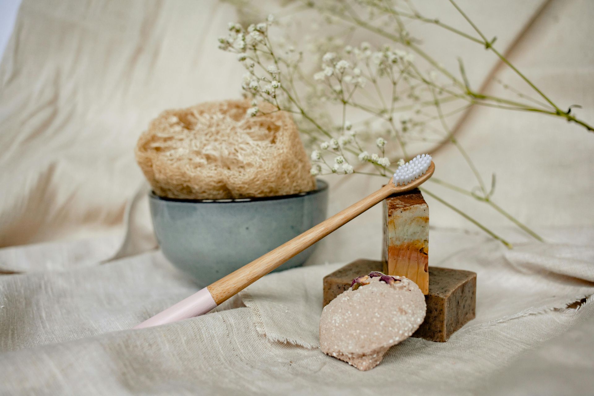 A bamboo toothbrush, a loofah in a gray bowl, and stacked bars of soap on a beige fabric background with white flowers.