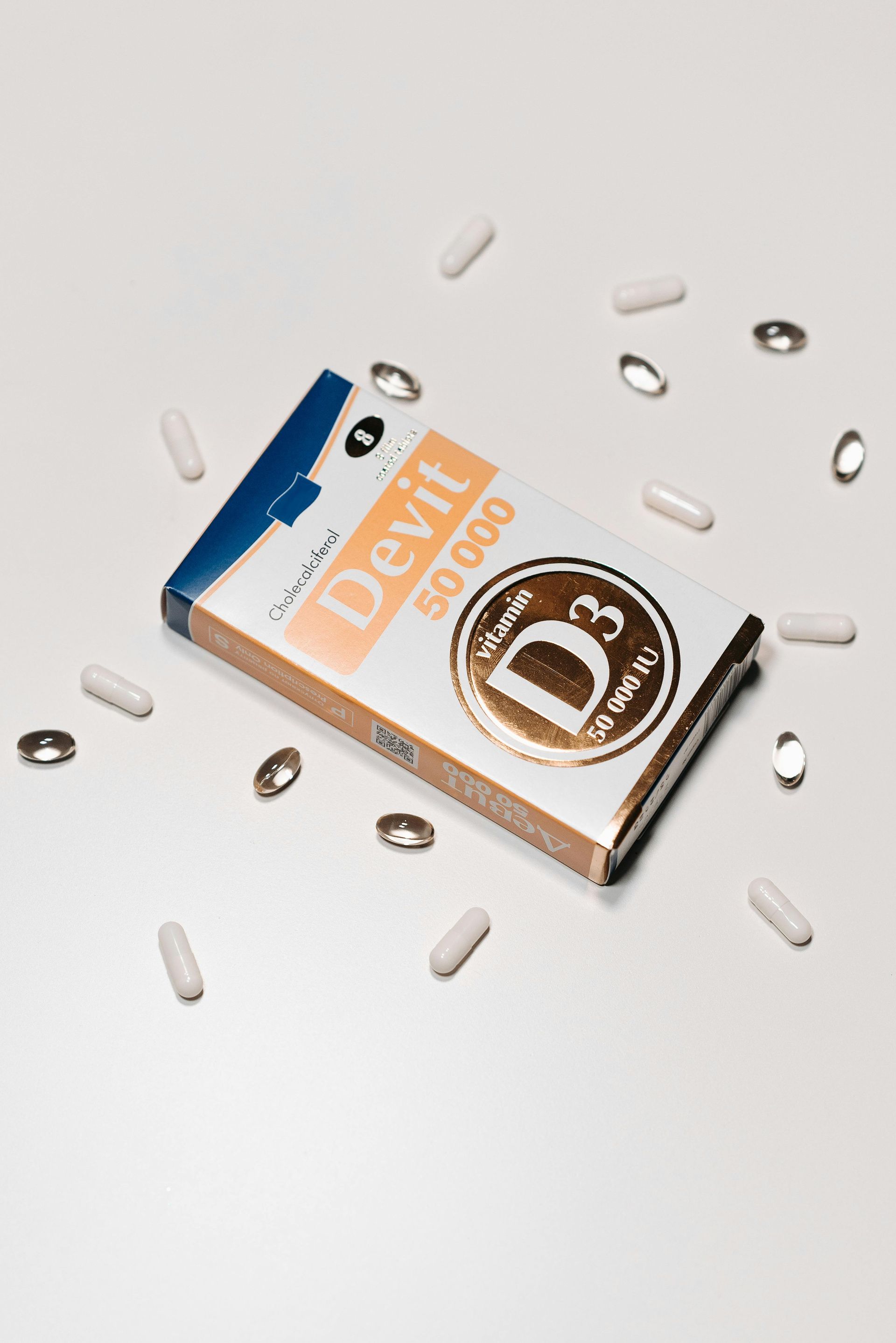 A box of Devit 50,000 vitamin D3 supplements surrounded by several loose capsule and pill supplements on a white surface.