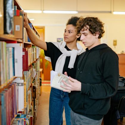 Two people stand in a library aisle, one reaching for a book on a wooden shelf while the other holds a book.