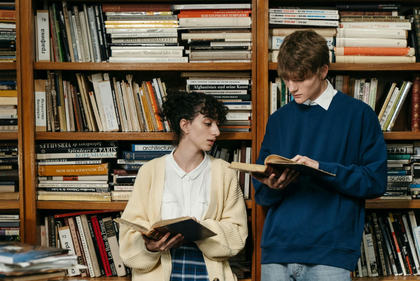 Two people read books while standing in front of a tall, packed wooden bookshelf.