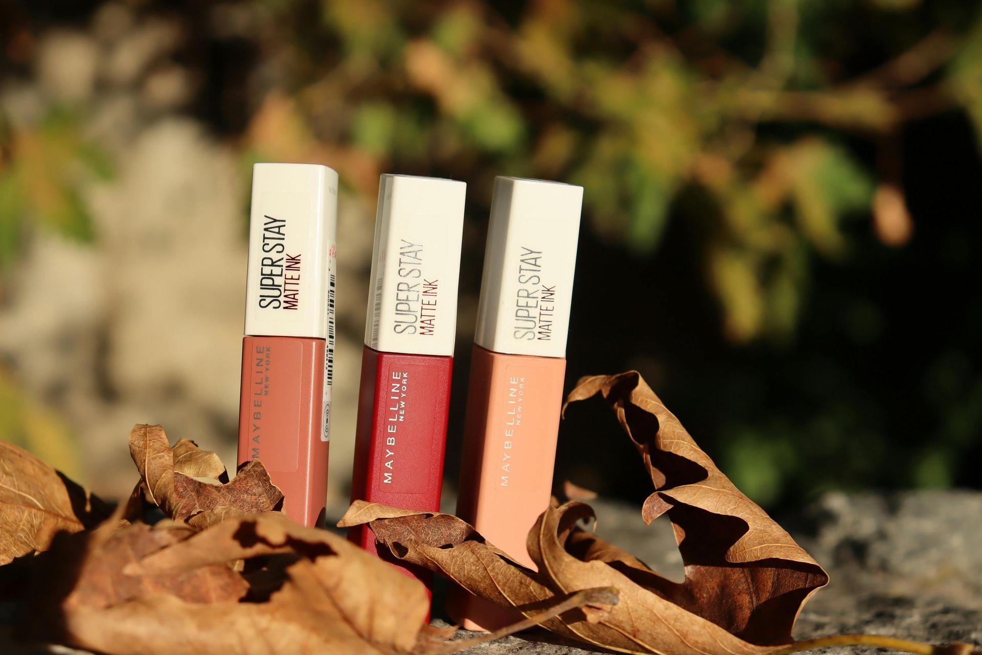 Three Maybelline SuperStay Matte Ink lipsticks in nude, pink, and peach shades arranged on dry leaves outdoors.