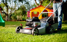 Person pushing a gray lawn mower across a green yard, with a playhouse in the background