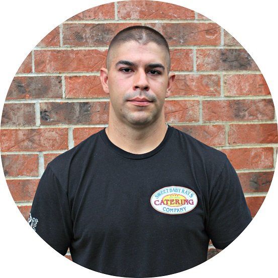 A man wearing a catering company shirt stands in front of a brick wall