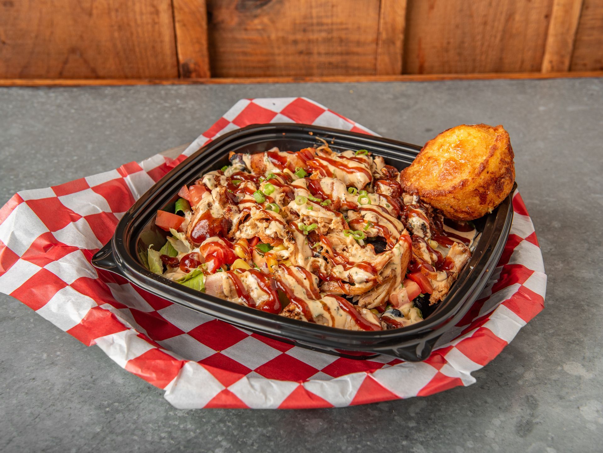 A chicken sandwich with lettuce and tomato next to a black bowl of baked beans on a red and white gingham parchment paper