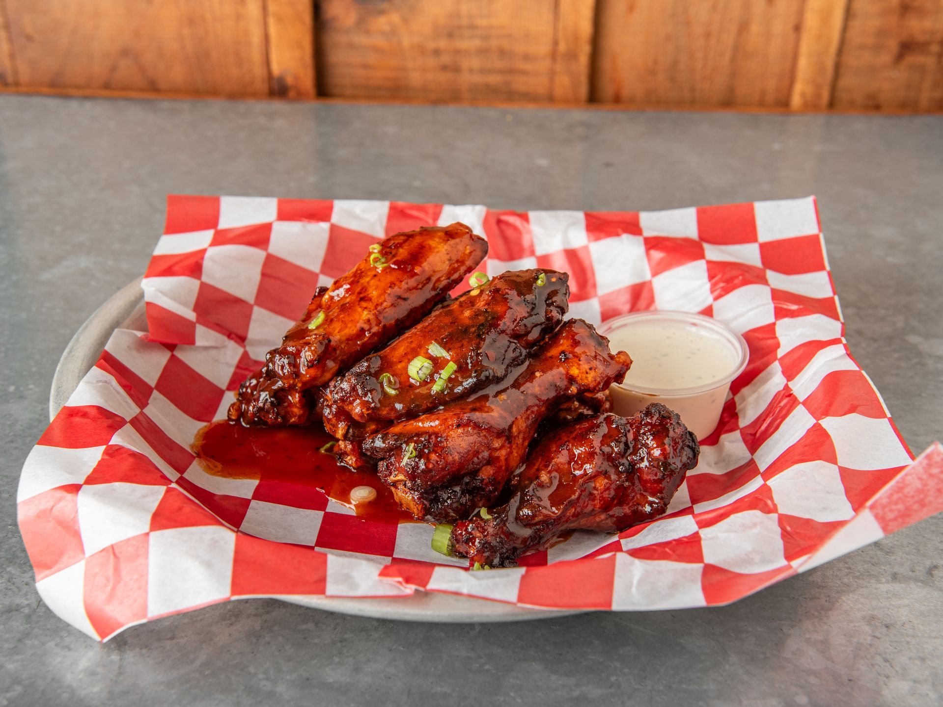 A stack of barbecue chicken wings with ranch dressing on a red and white gingham parchment paper