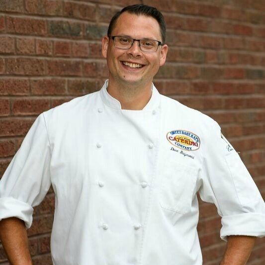 A man in a chef 's uniform is smiling in front of a brick wall