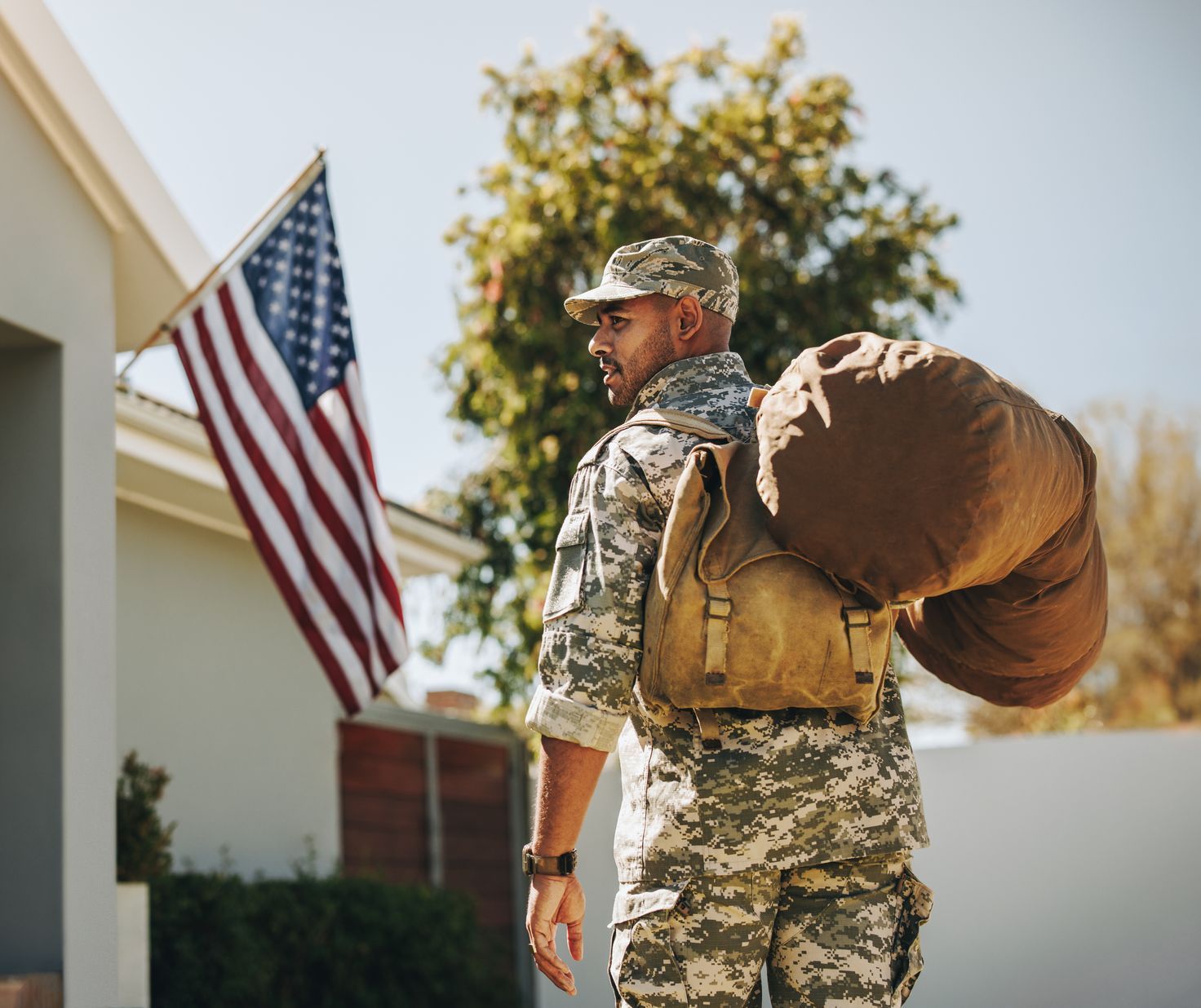 A soldier with a backpack is walking towards a house with an american flag in the background.