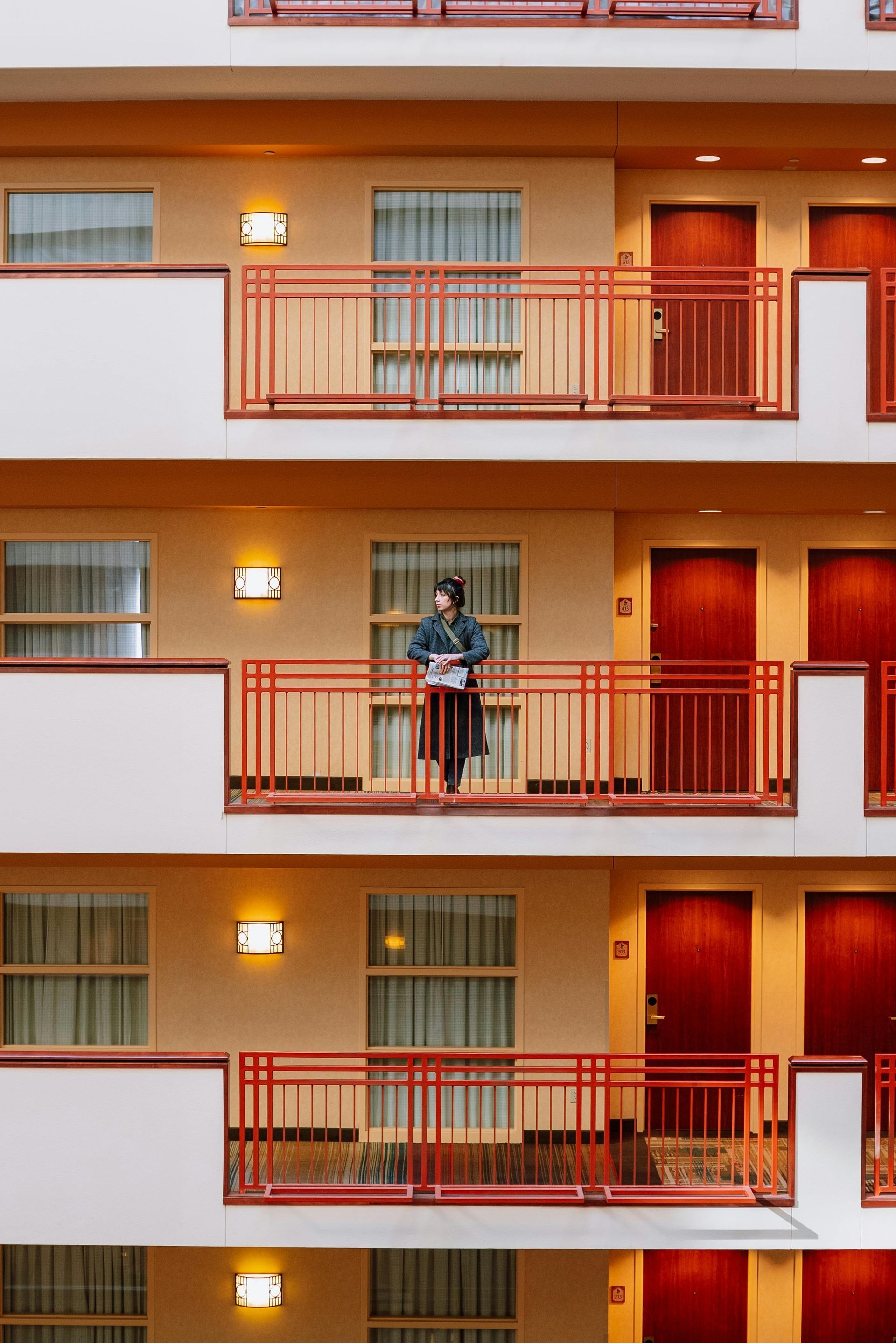 Person standing on a hotel balcony holding a basket. Red railing, multiple floors with doors and windows.