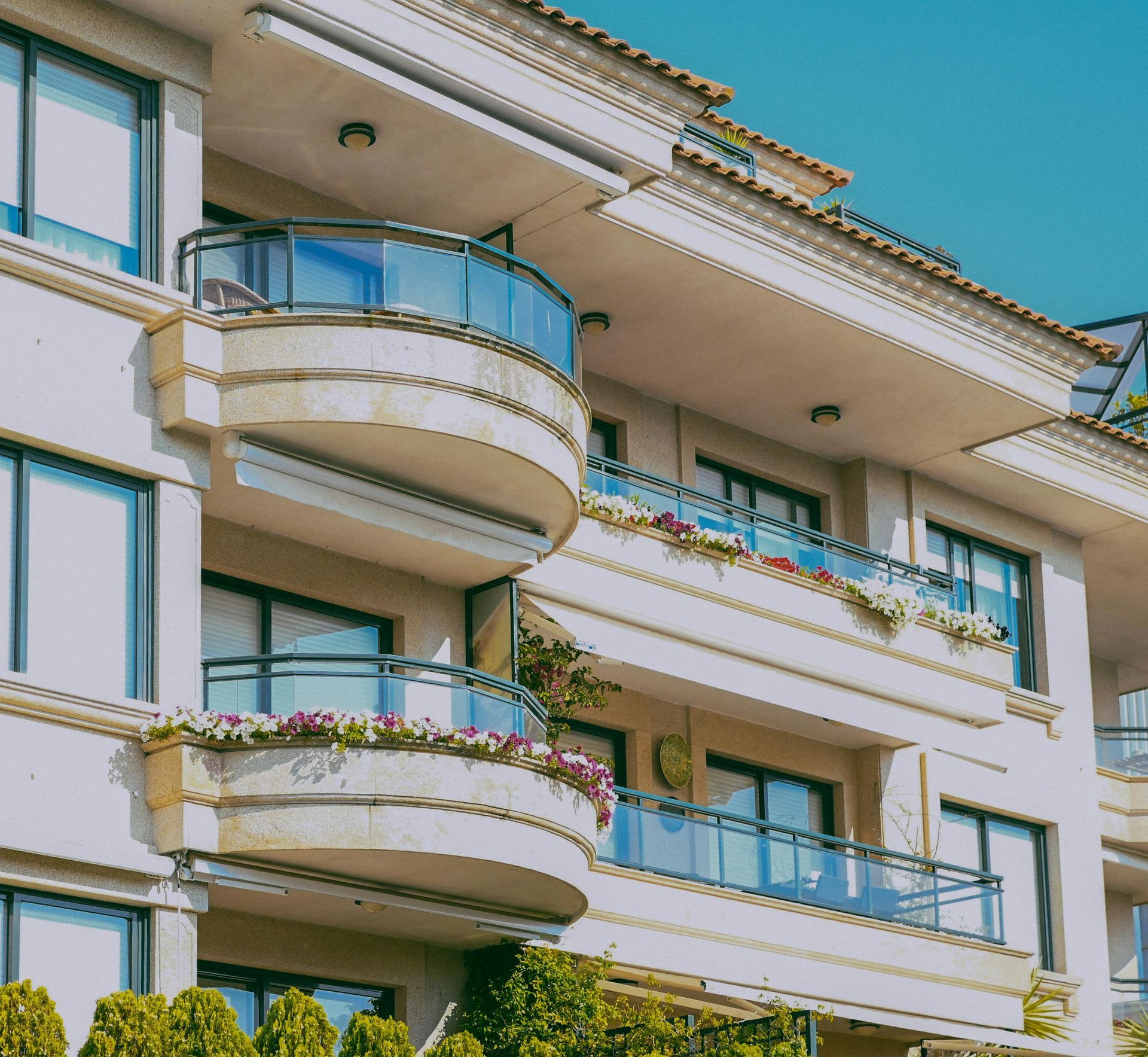 A multi-story beige apartment building featuring curved balconies with glass railings and planters filled with flowers.