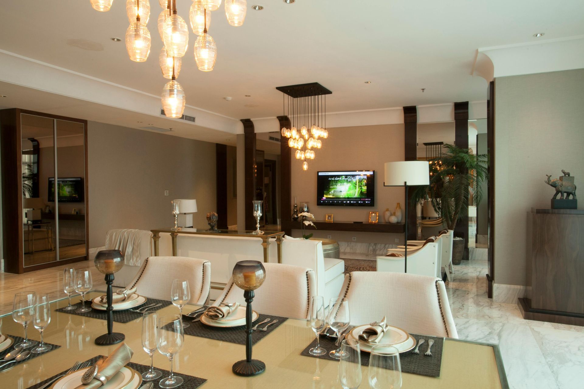 A dining room flows into an elegant living area with two modern chandeliers, neutral furniture, and a wall-mounted TV.