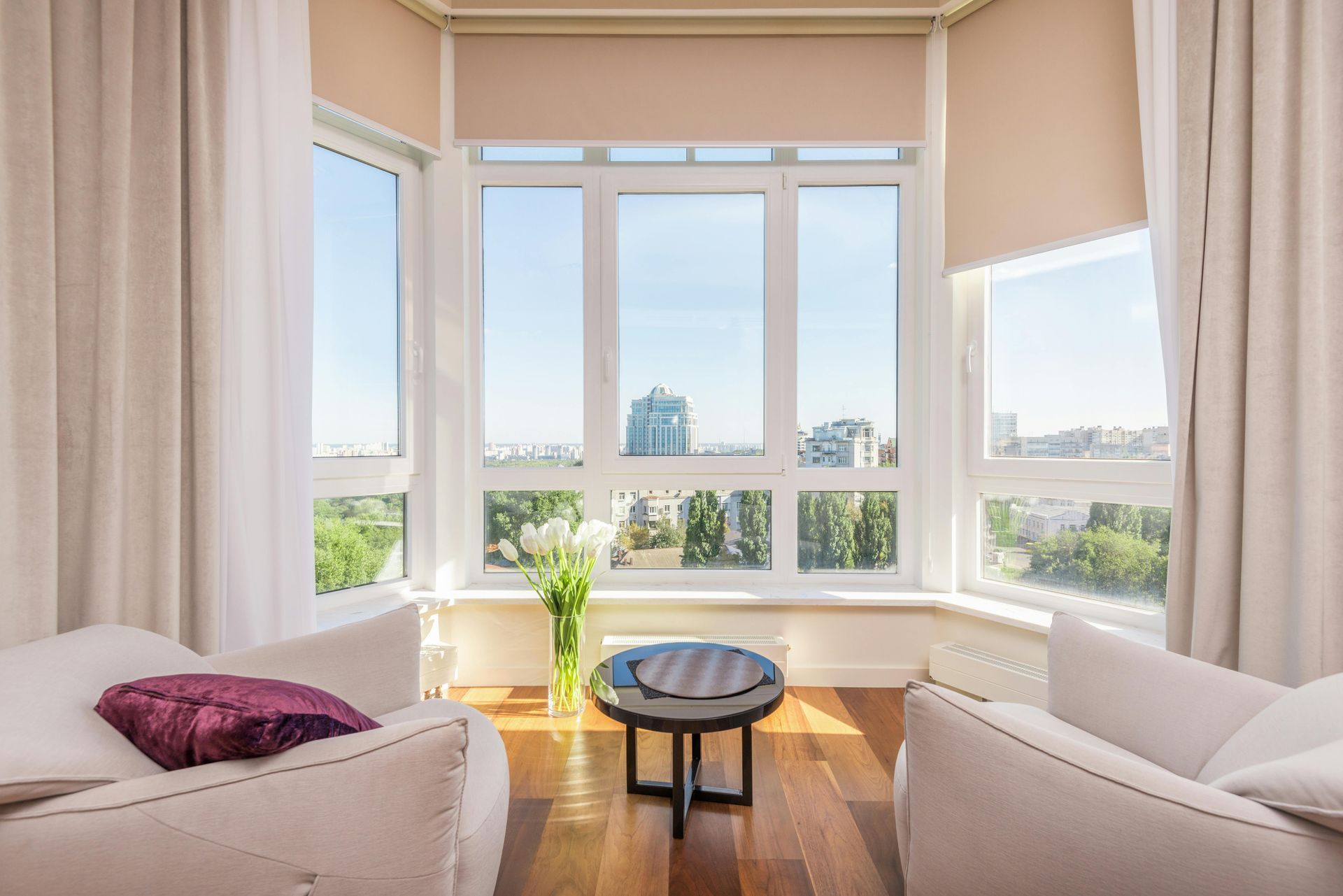 Bright room with white chairs, small table, large windows with city view, white curtains.