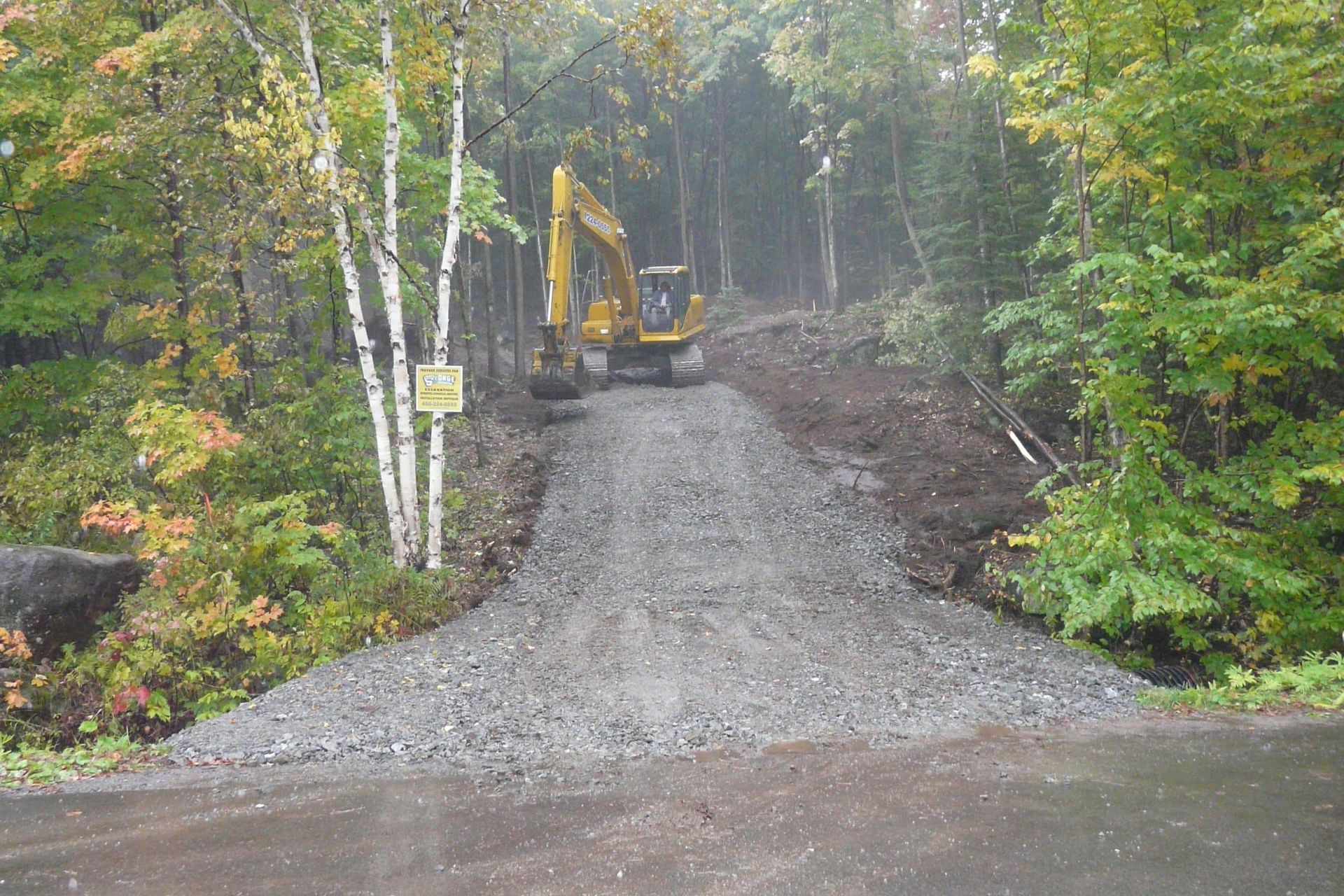 Une pelleteuse jaune trône au sommet d'une allée de gravier fraîchement aménagée qui mène à une zone boisée.